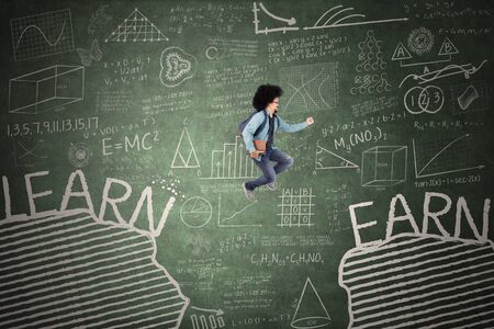 Male college student jumps between gap with words of learn and earn while holding a bookの写真素材