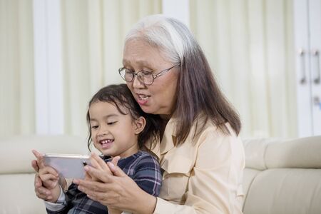 Old woman using a mobile phone with her grandchild at home while sitting on the sofa
