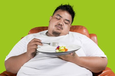 Lose weight concept. Obese man looks lazy to eat a plate of healthy food on the sofa and sitting in the studioの写真素材