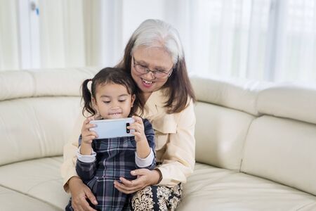Picture of cute little girl using a mobile phone with her grandmother at home while sitting on the...