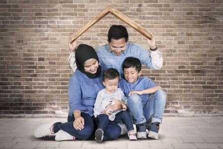 Young Muslim family holding a house roof symbol from cardboard over their heads while playing together in bricks wall backgroundの写真素材