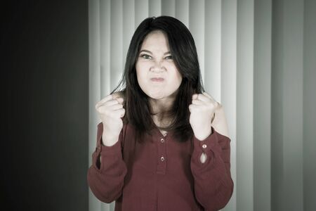 Picture of young Asian woman looks angry while clenching her fists near the windowの写真素材