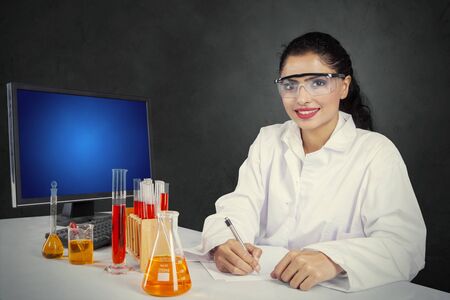 Beautiful female scientist smiling at the camera while sitting with test tubes in the studioの写真素材