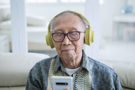 Close up of old man enjoying music with a mobile phone and headphones in the living room at homeの写真素材