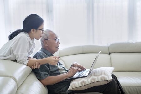 Picture of old man using a laptop computer with his daughter while sitting on the sofa. Shot at homeの写真素材