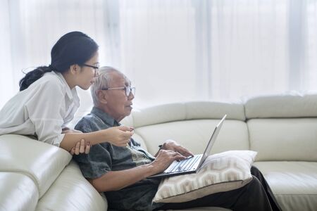 Picture of old man using a laptop computer with his daughter while sitting on the sofa. Shot at homeの素材 [FY310127963889]