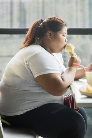 Side view of an overweight woman eating an ice cream cone and sitting in the cafeの写真素材