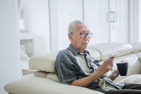 Image of an old man enjoying leisure time by hot tea and mobile phone at homeの写真素材