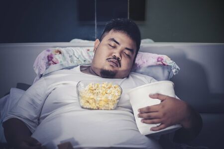 Unhealthy lifestyle concept: Young fat man sleeping with junk foods on the bedの写真素材