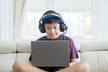 Picture of Asian preteen boy sitting on the couch while using a laptop and headphones at homeの写真素材