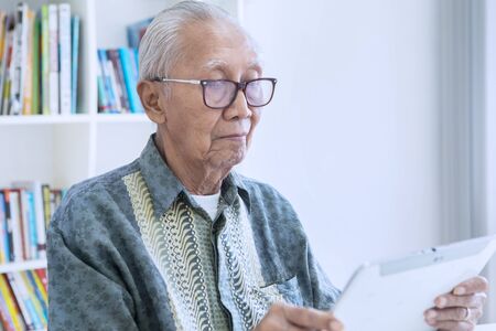 Senior man using a digital tablet while wearing glasses in the library with bookcase background