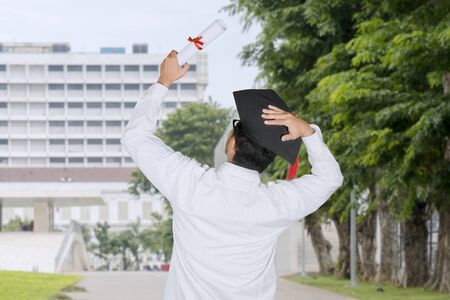 Back view of a young man celebrating his graduation by lifting a diploma at outdoorsの写真素材