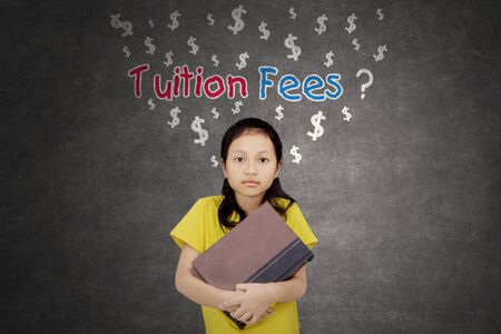 Pensive schoolgirl holding books while standing with a question of tuition fees on a blackboardの写真素材