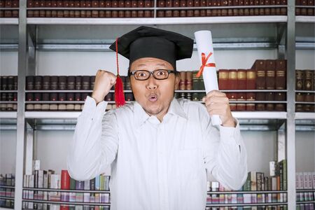 Picture of a happy man celebrating his graduation by raising hands and diploma while standing in the...