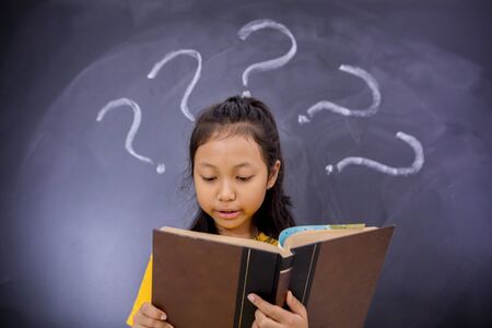 Nerdy schoolgirl reading a book while standing with question marks in the classroom