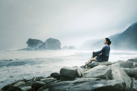 Beautiful woman sitting on the stone while enjoying a foggy Papuma beach view at morning timeの写真素材