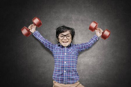 Picture of a little boy looks happy while lifting two dumbbells with blackboard backgroundの写真素材