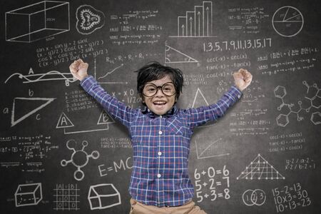 Happy little boy raising hands and jumping in the classroom with doodles backgroundの写真素材