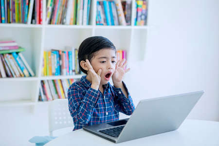 Picture of surprised little boy using a laptop computer while sitting in the library with bookcase backgroundの写真素材