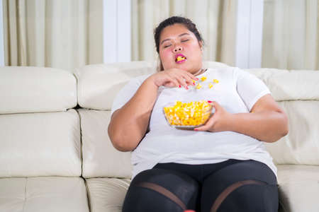 Fat woman falling asleep during eating a bowl of popcorn and sitting on the couch. Shot at homeの写真素材