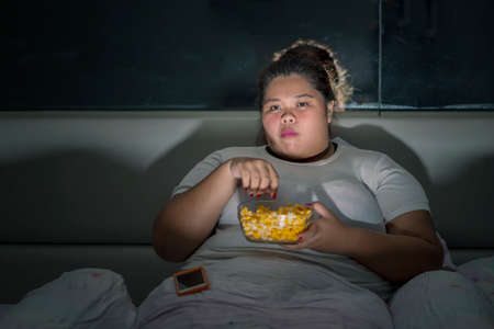 Overweight woman enjoying a bowl of popcorn while watching television in the bedroom. Unhealthy lifestyle conceptの写真素材