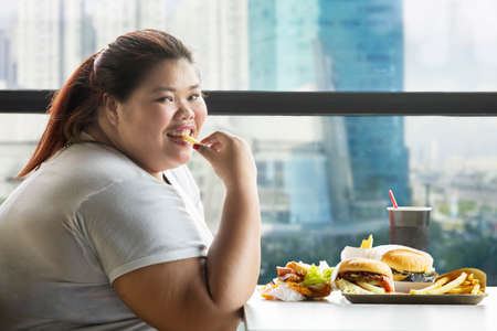 Obese woman smiling at the camera while eating french fries and hamburgers in the cafeの写真素材