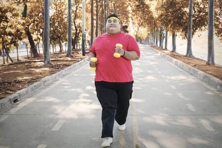 Attractive overweight man running at the autumn park while carrying dumbbells on the morning. Shot outdoorsの写真素材