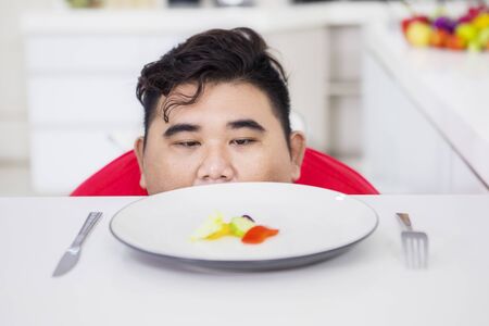 Picture of Asian fat man peeking out of the table while looking at salad on a plate