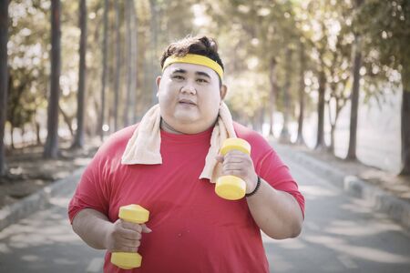 Young obese man holding two dumbbells while jogging in the park at autumn timeの写真素材