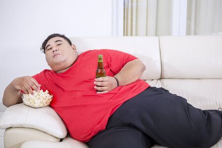 Picture of overweight man watching TV while eating popcorn and drinking beer on the couchの写真素材