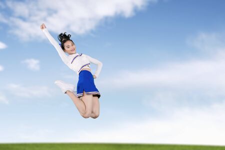 Portrait of Asian cheerleader girl smiling at the camera while jumping in the field with blue sky backgroundの写真素材