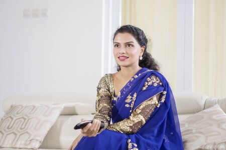 Young Indian woman wearing blue saree clothes while watching TV in the living room. Shot at home