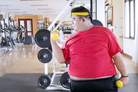 Picture of young obese man exercising his biceps with two dumbbells in the gym centerの写真素材