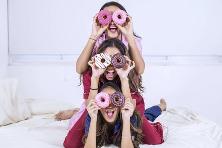 Group of happy teenage girls covering their eyes with donuts while having fun in the bedroomの写真素材