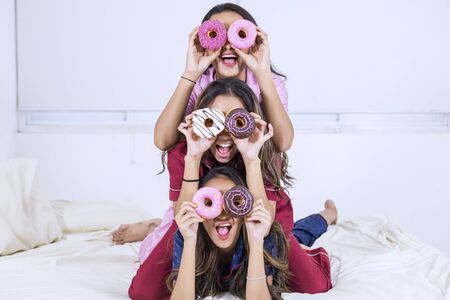 Group of happy teenage girls covering their eyes with donuts while having fun in the bedroomの素材 [FY310131354583]