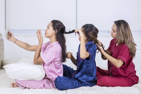 Group of pretty teenage girls tying hair to each other while taking a selfie photo with mobile phone in the bedroom at homeの写真素材