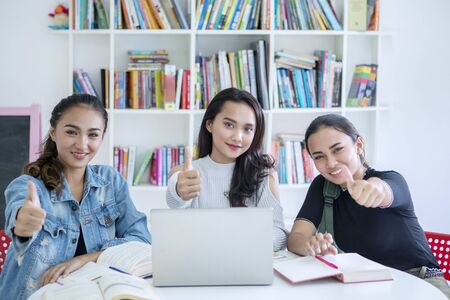 Group of pretty teenage girls showing thumbs up at the camera while studying together in the libraryの写真素材