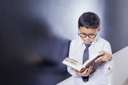 Picture of schoolboy reading a book while leaning on the blackboard in the classroomの写真素材