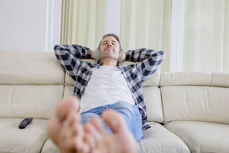 Mature man enjoying music with headphones in the living room while relaxing on the sofa at homeの写真素材