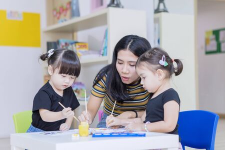 Young woman teaching two children to painting on a paper while playing in the kindergartenの写真素材