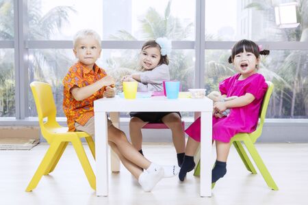 Group of little children having lunch together while sitting near the windowの写真素材