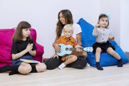 Nursery teacher helping a little boy to play the guitar and sitting with classmate in the kindergartenの写真素材