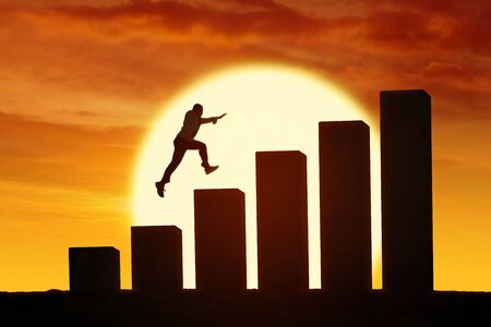 Silhouette of unidentified businessman jumping and climbing through giant business charts out in somewhere with sun backgroundの写真素材