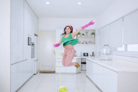 Beautiful Asian woman jumping happily and joyfully while sweeping the floors in the kitchenの写真素材
