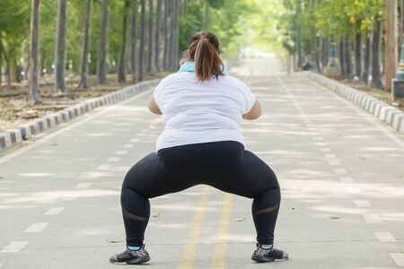 Back view of unidentified fat woman wearing sportswear while squatting and stretching her thighs in...