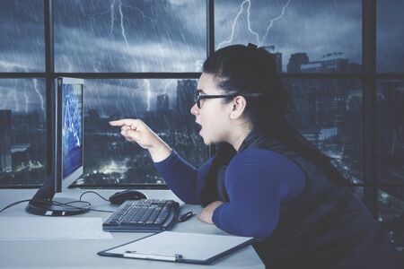 Portrait of beautiful Asian businesswoman wearing formal attire while feeling annoyed at the business chart at her desktop in her office with stormy city backgroundの写真素材