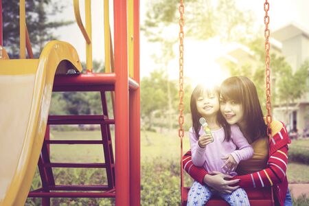 Beautiful Asian woman smiling at camera with her daughter while holding her daughter on a swing at the parkの写真素材