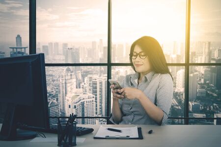 Portrait of beautiful Asian businesswoman texting someone while working on her desktop in her office at sunny day with cityscape backgroundの写真素材
