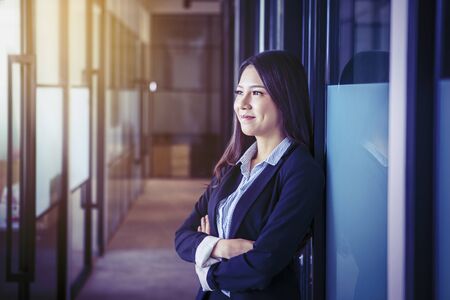 Portrait of beautiful Asian businesswoman wearing a suit while staring at the sunlight with a smile on the office hallwayの写真素材