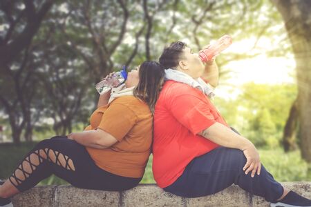 Fat Asian couple chugging water from their bottles, while taking a break after working out on the park pavements at sunny dayの写真素材